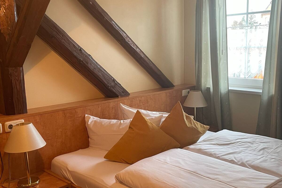 Een hotelkamer met een houten plafond, een bed met witte lakens en twee kussens, een nachtkastje met een lamp en een raam met gordijnen.