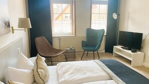 Hotelkamer met een bed, twee stoelen, een kleine tafel, een tv op een standaard en ramen met gordijnen.