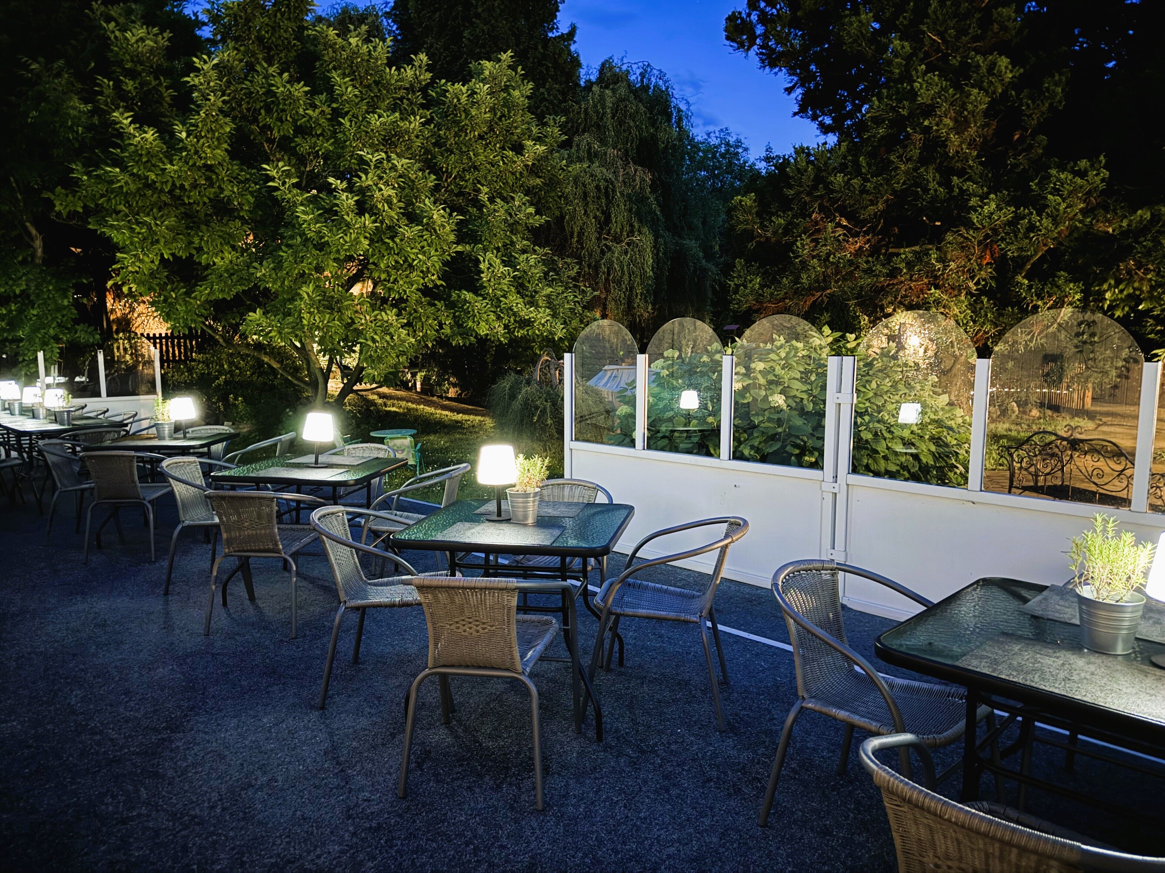 Un patio exterior con mesas y sillas, rodeado de árboles y una valla blanca.