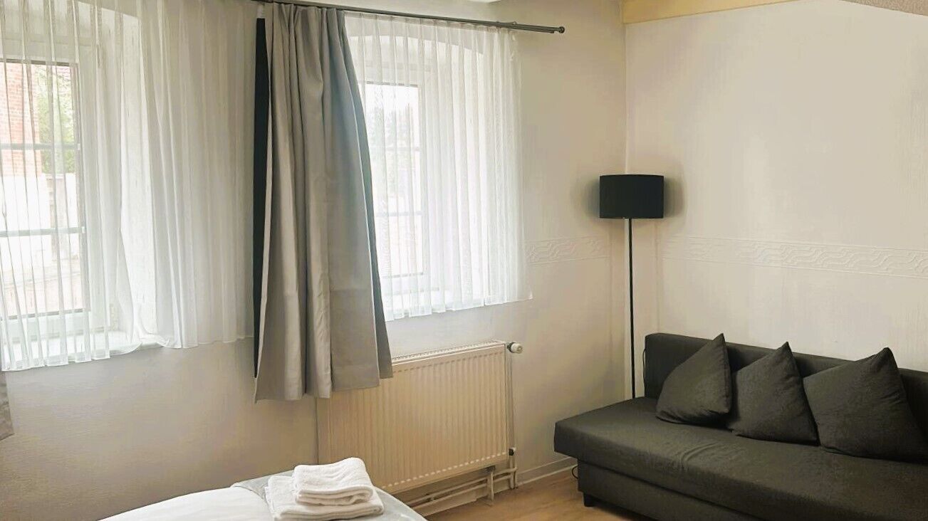 Una habitación con una cama, un sofá oscuro, una lámpara de pie y una ventana con cortinas traslúcidas.