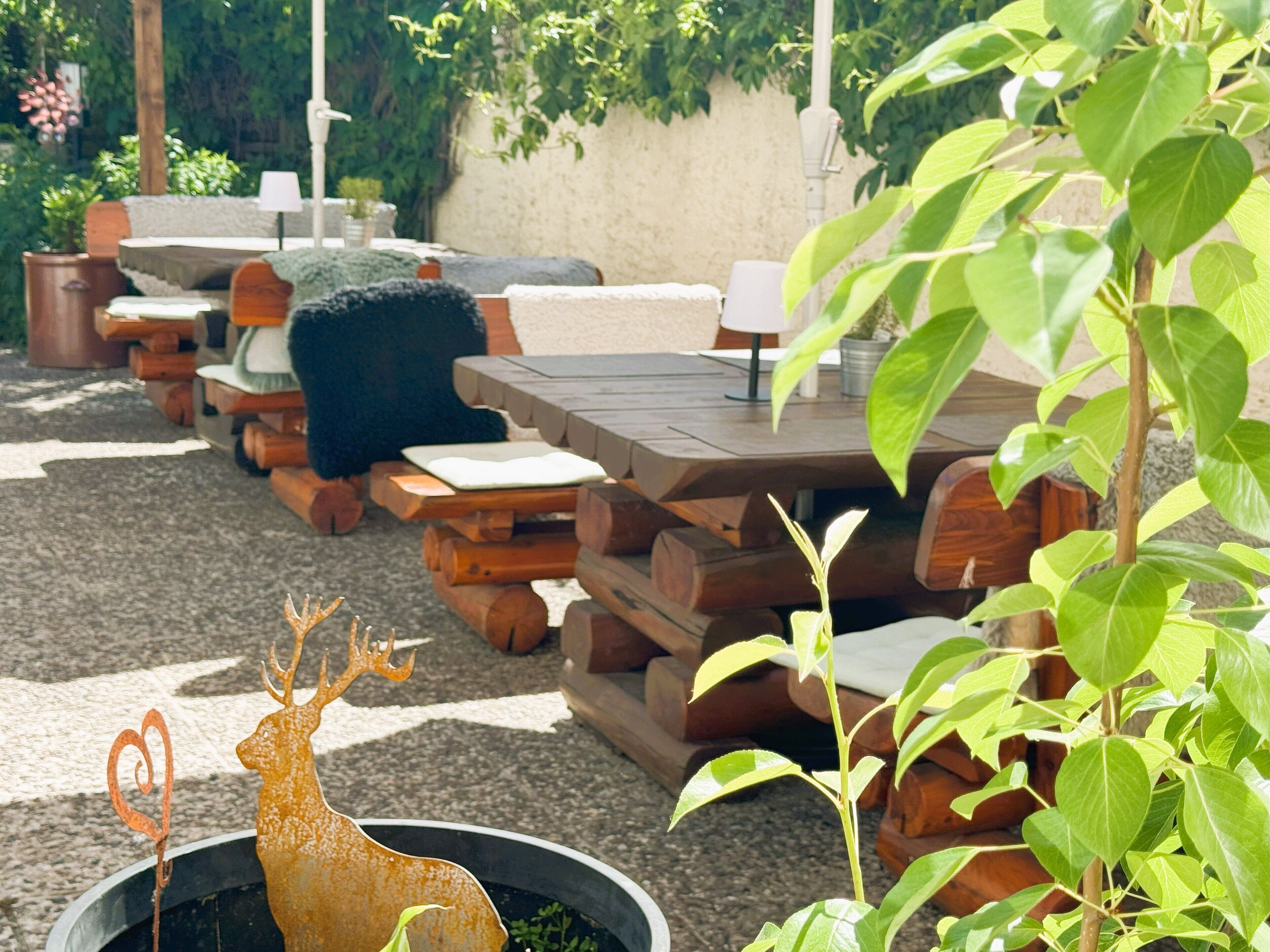 An outdoor seating area with wooden tables and benches, surrounded by greenery and a decorative deer sculpture.
