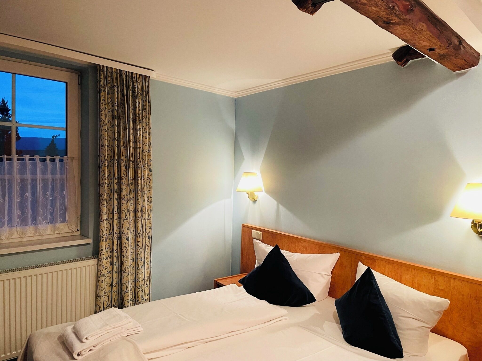 Ein ordentlich bezogenes Bett mit weißen Laken und dunklen Kissen, ein Fenster mit gemusterten Vorhängen und ein Holzkopfstück.
