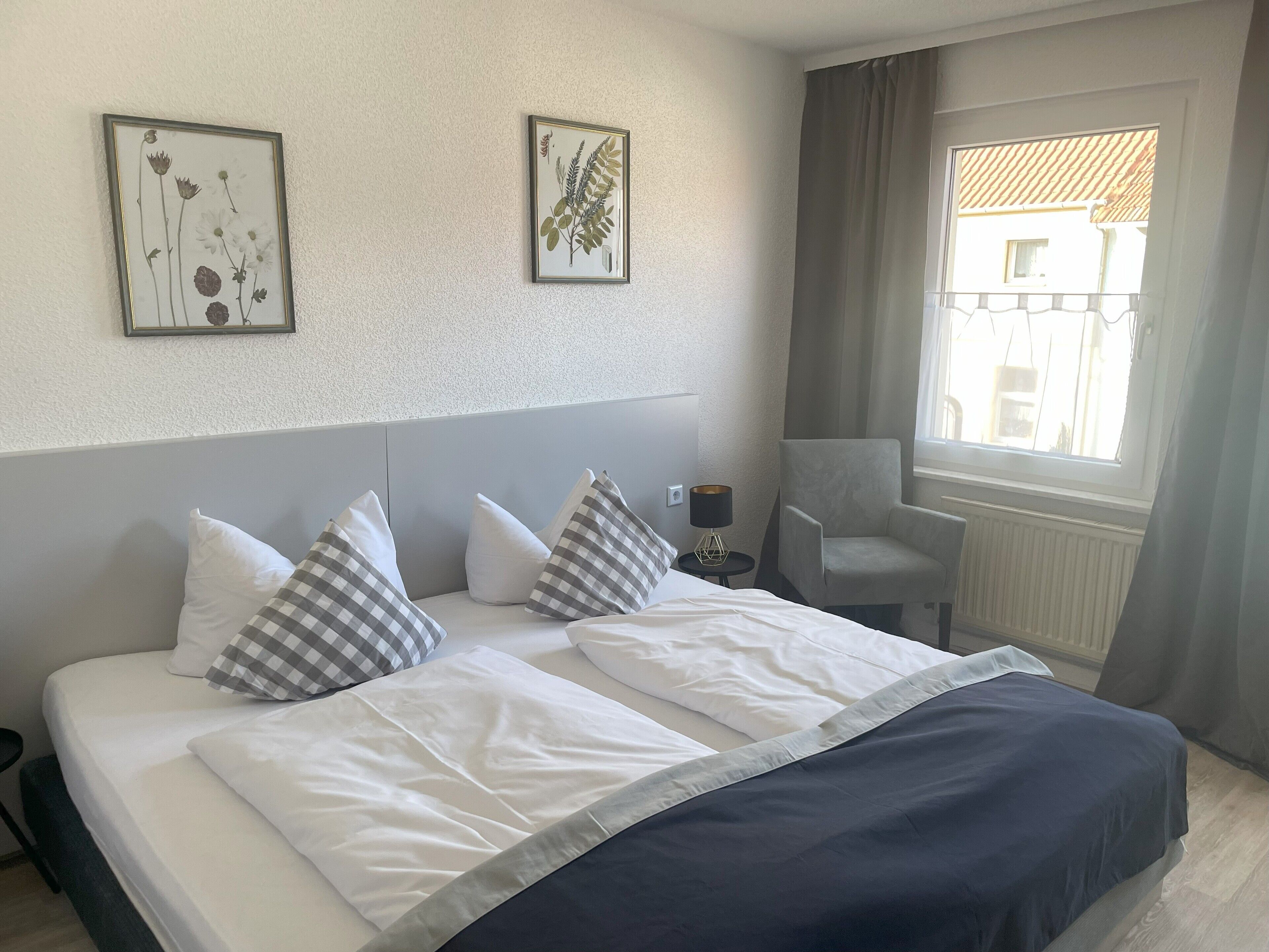 Camera d'albergo con un letto, due cuscini, una sedia, un comodino e due stampe incorniciate a tema botanico.