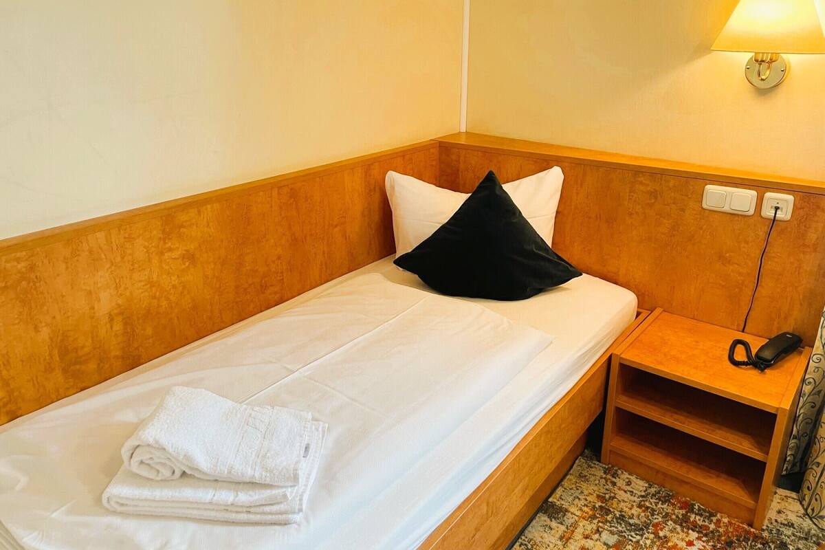 Een eenpersoonsbed met witte lakens en een zwart kussen, een nachtkastje met een lamp en een telefoon.