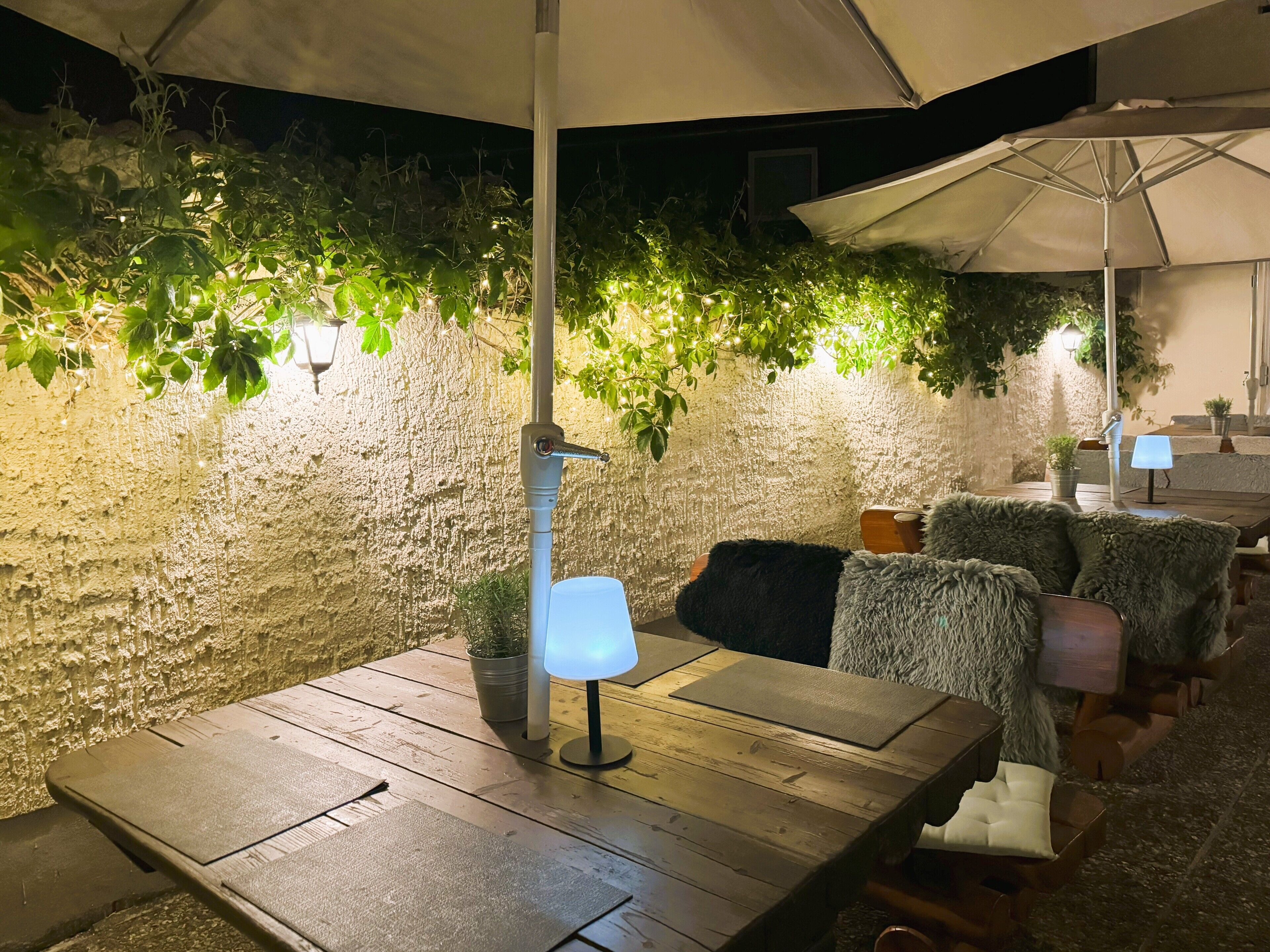 Un área de estar al aire libre con mesas y sillas de madera, una pared con textura y plantas en macetas.