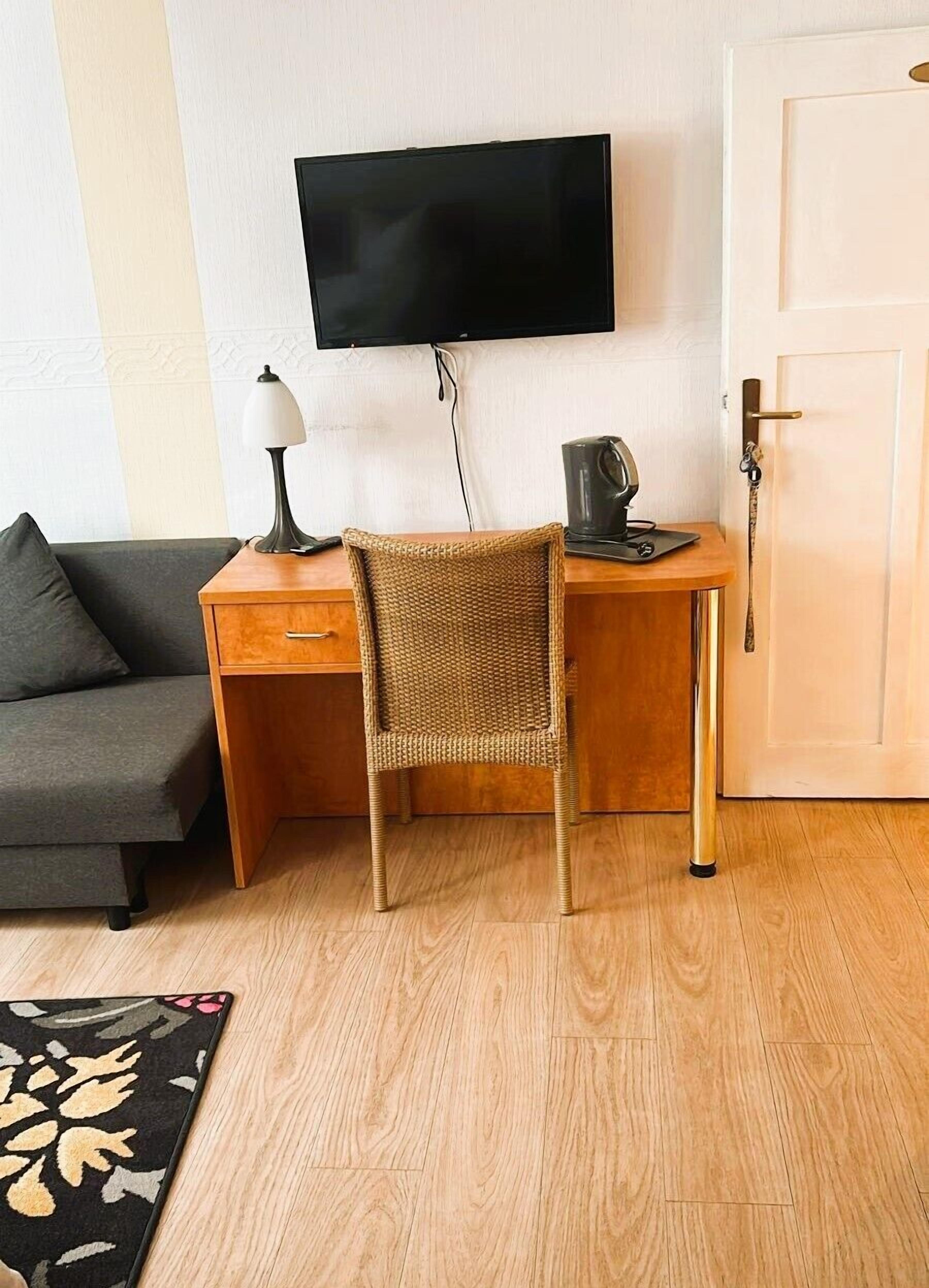 Une pièce comprenant un bureau en bois, une chaise en osier, un canapé gris, un téléviseur à écran plat, une lampe et une machine à café.