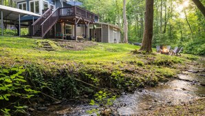 A cabin with a deck, a stream, and outdoor seating by the fire. - Hoopers Creek Cottage | Creekside Retreat Near Asheville + Hendersonville (Fletcher)