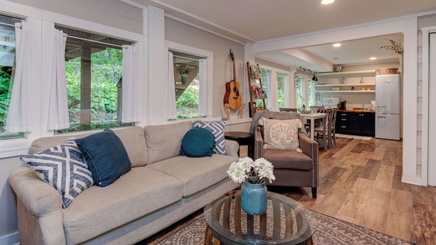A spacious living room with a beige sofa, a coffee table with a vase, and a dining area with a table and chairs. - Hoopers Creek Cottage | Creekside Retreat Near Asheville + Hendersonville (Fletcher)