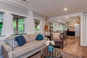 A spacious living room with a beige sofa, a coffee table with a vase, and a dining area with a table and chairs. - Hoopers Creek Cottage | Creekside Retreat Near Asheville + Hendersonville (Fletcher)