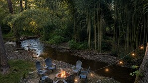 A campsite with a fire pit and chairs by a stream in a forest. - Hoopers Creek Cottage | Creekside Retreat Near Asheville + Hendersonville (Fletcher)