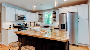 A modern kitchen with white cabinetry, stainless steel appliances, a large island with stools, pendant lighting, and a window providing natural light. - Hoopers Creek Cottage | Creekside Retreat Near Asheville + Hendersonville (Fletcher)
