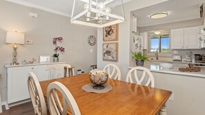 Un comedor con mesa de madera, sillas blancas, una araña y una cocina al fondo.
