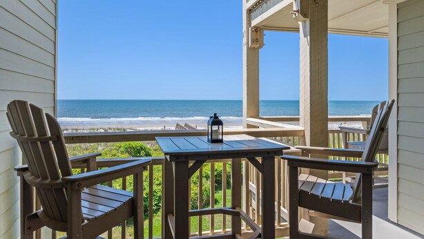 Una terraza de madera con dos sillas Adirondack y una mesa, con vista a una playa y al océano.