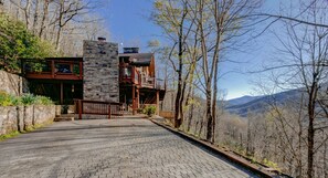 A stone and wood cabin with a balcony, surrounded by trees and a mountain view. - Falling Water Lodge | Gorgeous 5BR Mountaintop Lodge (Burnsville)