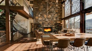 A spacious living area with a stone fireplace, a dining table, and a staircase leading to an upper level.