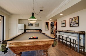 A pool table with a wooden finish, a hanging green pendant light, and a bar area with stools. - Falling Water Lodge | Gorgeous 5BR Mountaintop Lodge (Burnsville)