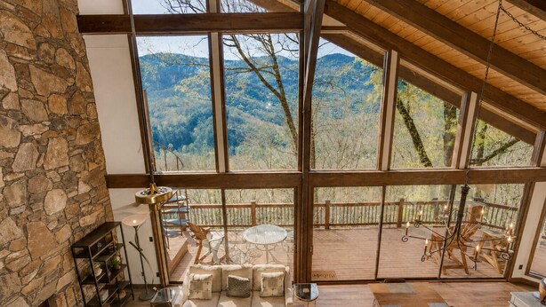 A spacious room with a stone fireplace, large windows offering mountain views, a cozy seating area, and a wooden deck.