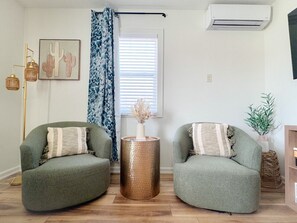 A living room with two green armchairs, a round copper side table, a cactus artwork, a window with blinds, a blue patterned curtain, and an air conditioning unit.