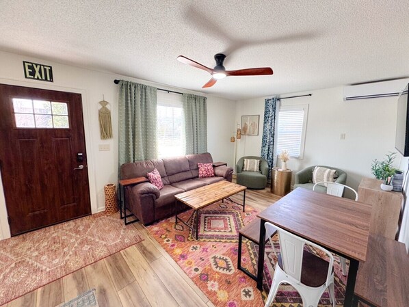 A living room with a sofa, coffee table, dining table, and chairs. There is a ceiling fan, a door with an 'EXIT' sign, and a window with curtains.