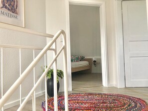 Un escalier avec une rampe blanche, un tapis coloré à motifs au pied de l’escalier, un lit avec un coussin à fleurs et une œuvre d’art encadrée accrochée au mur.