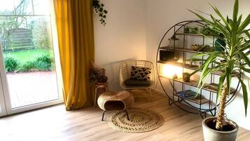 Ein Zimmer mit großem Fenster, gelbem Vorhang, Korbsessel, rundem Teppich, Blumentopf und einer Metallregaleinheit mit Büchern und Dekoration.