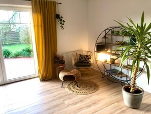 A room with a large window, a yellow curtain, a wicker chair, a round woven rug, a potted plant, and a metal shelving unit with books and decorative items.