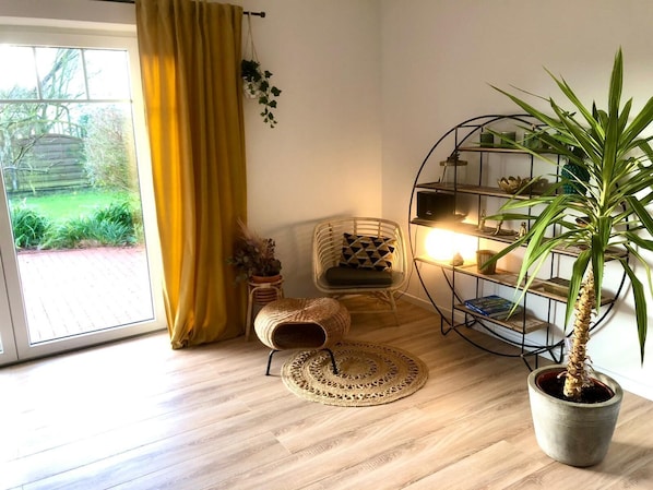 A room with a large window, a yellow curtain, a wicker chair, a round woven rug, a potted plant, and a metal shelving unit with books and decorative items.