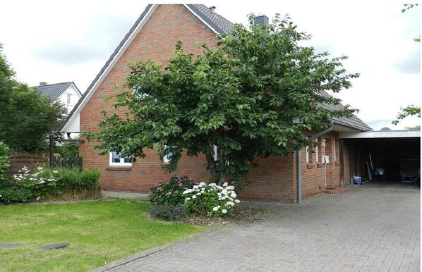 A brick building with a triangular roof, a large tree in front, and a paved driveway.