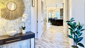 A hallway with a marble wall, a round mirror, a white bowl, a potted plant, and a kitchen area in the distance. - Golfers` Paradise. Large Townhouse By 1st Hole In Private Gated Community (Gulf Shores)