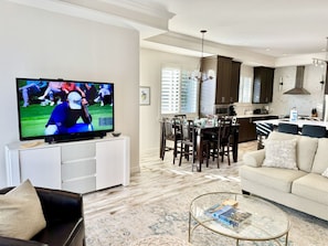 Una sala moderna con un televisor de pantalla plana, una zona de comedor con mesa y sillas, una cocina con armarios oscuros y un sofá color beige.
