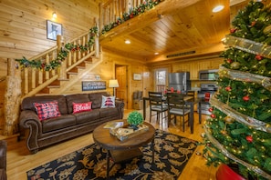 A cozy log cabin interior with a wooden staircase, a living room with a brown leather sofa, a dining area, and a decorated Christmas tree.
