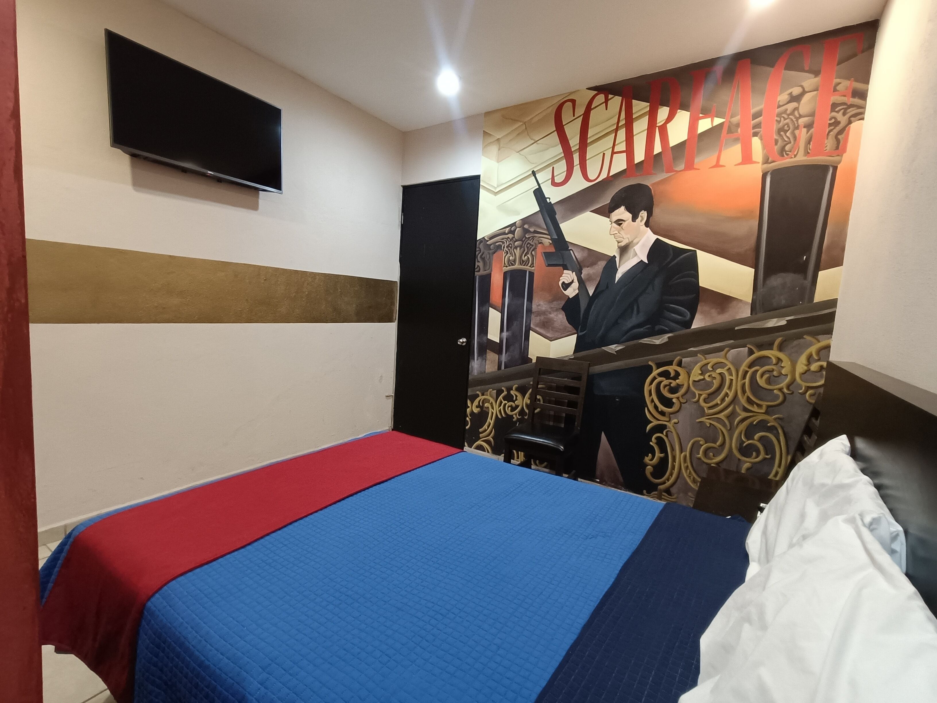 A hotel room with a bed, a television mounted on the wall, and a mural with the word 'SCARFACE' and a figure holding a gun.