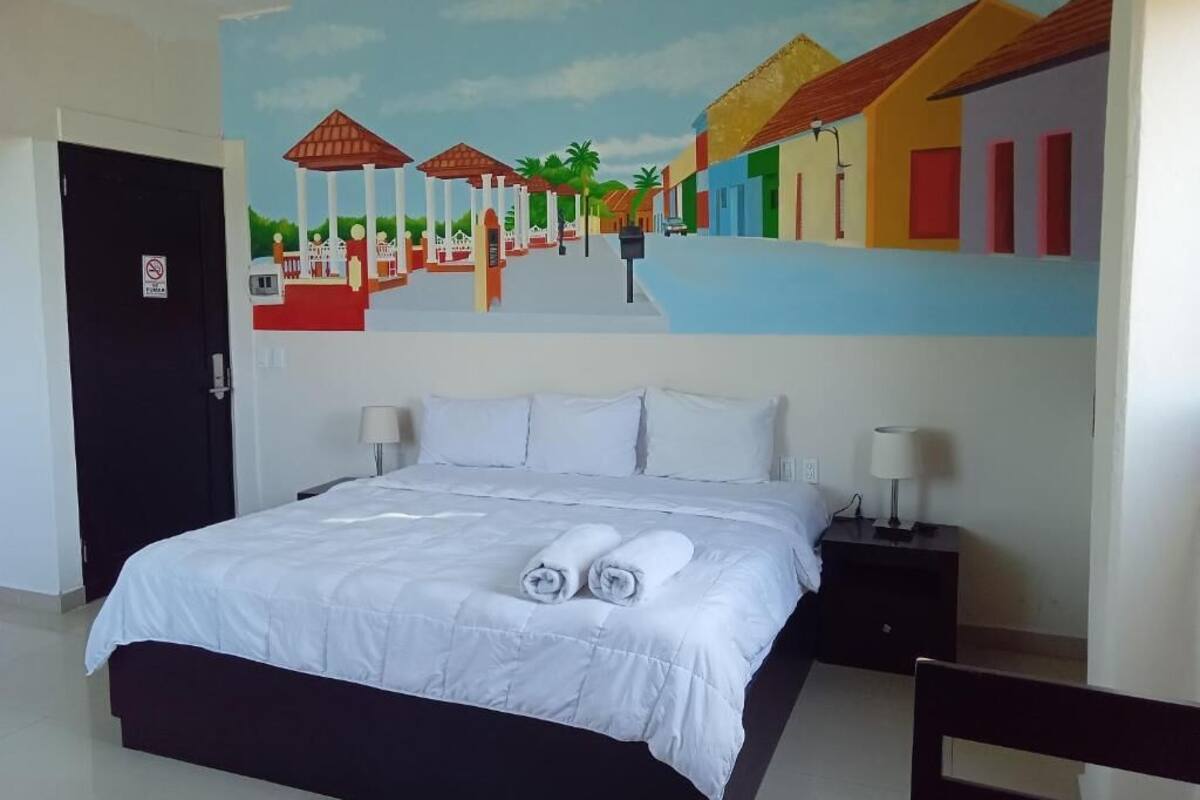 A hotel room with a bed, two bedside tables, a painting of a street scene, and a door with a 'No Entry' sign.