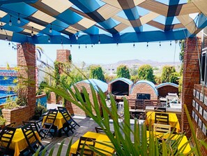 Eine Terrasse im Freien mit Backöfen, gelben Tischen und Stühlen unter einem blau-beigen Dach.