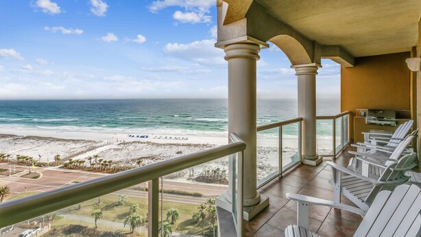 A balcony with a view of the beach and ocean. There are white chairs and a grill on the balcony.