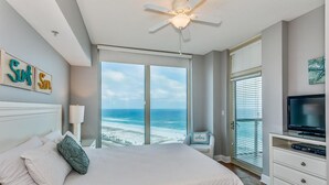 A bedroom with a large bed, a ceiling fan, a TV, and a view of the ocean.