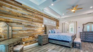 A bedroom with a wooden accent wall, a bed with a blue quilt, a dresser, a bench, and a ceiling fan.