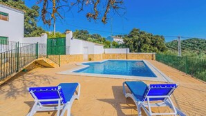 A pool with two blue lounge chairs, a stone wall, and a green fence.