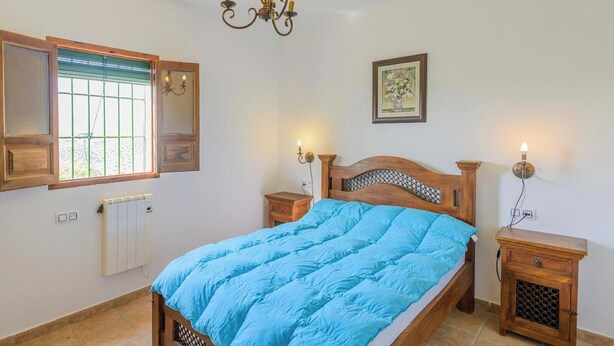 A bedroom with a wooden bed, a radiator, a window with blinds, bedside tables, and a wall-mounted light fixture.
