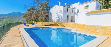 Una piscina con solárium de piedra, un edificio blanco con tejado de baldosas y vistas al paisaje.