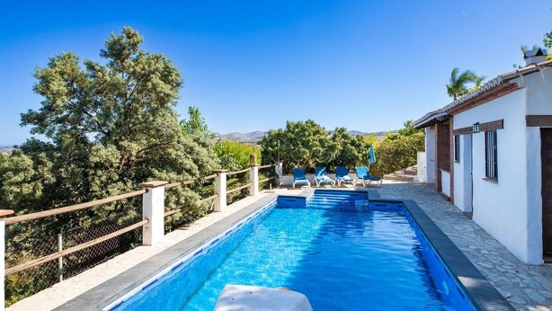 A rectangular swimming pool with a wooden deck and lounge chairs, surrounded by a white building with a tiled roof and a view of trees and hills.