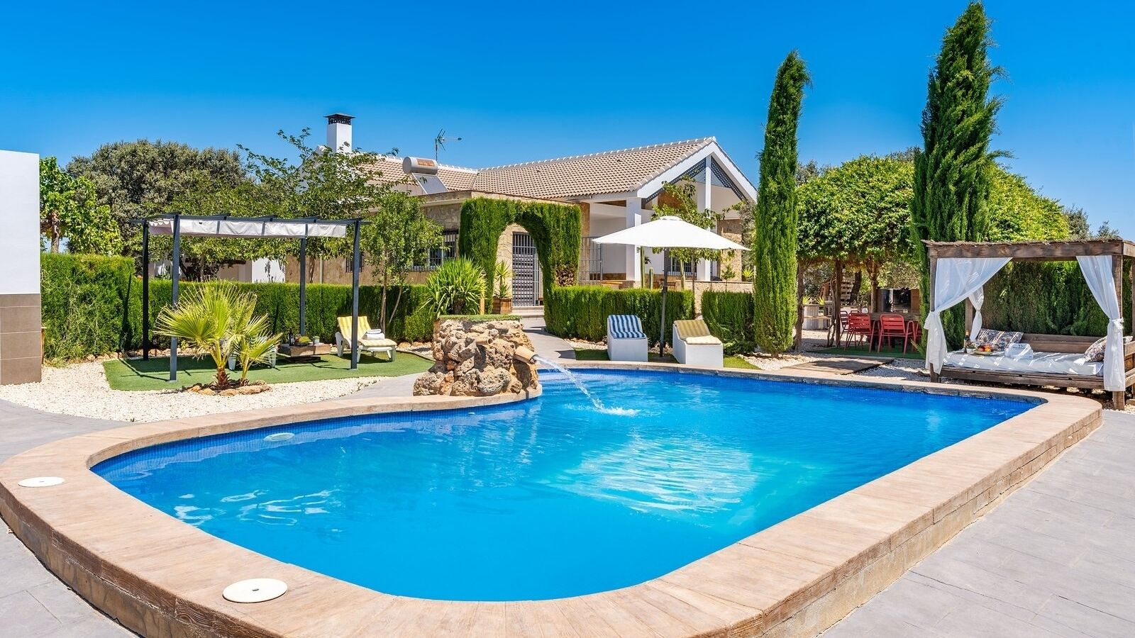 A swimming pool with a stone waterfall feature, surrounded by a well-maintained garden and a house with a tiled roof.
