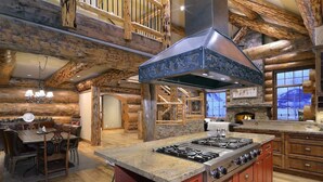 A rustic cabin interior with a large kitchen island, wooden beams, a fireplace, and a dining area. - Red Feather Ranch (Crested Butte)
