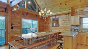 A wooden cabin interior with a large dining table, benches, a kitchen area, and a chandelier.