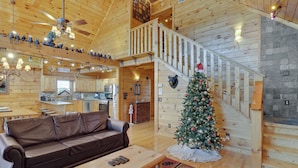A cozy cabin interior with a wooden staircase, a living room with a leather sofa and a coffee table, a kitchen area, and a decorated Christmas tree.