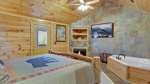 A log cabin interior with a wooden bed, a stone fireplace, a stone wall, a wooden bathtub, and a ceiling fan.