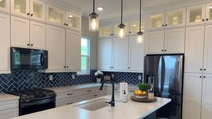 A modern kitchen with white cabinets, a black stove, a stainless steel refrigerator, and pendant lights.