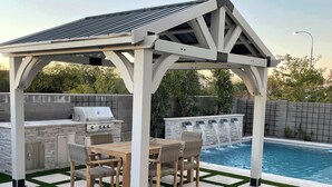 A covered outdoor patio with a grill, dining table, and chairs, adjacent to a swimming pool with a waterfall feature.