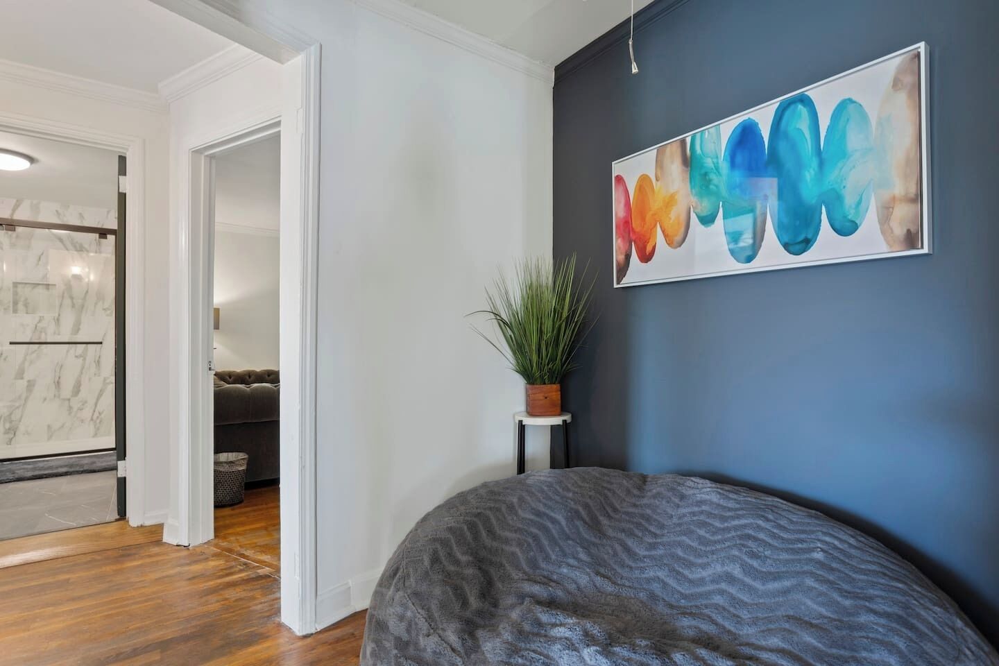 Quarto moderno com parede azul escuro, uma grande pintura abstrata, um bean bag cinza, uma pequena planta em vaso e área de chuveiro com padrão marmorizado.