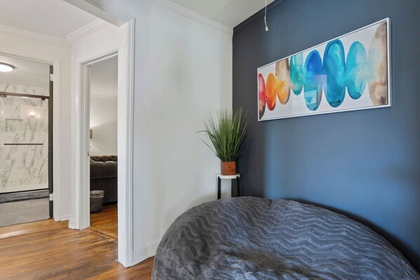 A modern bedroom with a dark blue accent wall, a large abstract painting, a grey bean bag, a small potted plant, and a marble-patterned shower area.